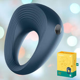 A blue, smooth, oval-shaped Satisfyer "Power Ring" Rechargeable Vibrating Cock Ring with a ridged surface and a metallic button on the side is displayed next to a yellow and green box that features an image of the device. The cock ring offers customizable vibration programs to boost stamina, set against a soft gradient background with blurred spots of light.